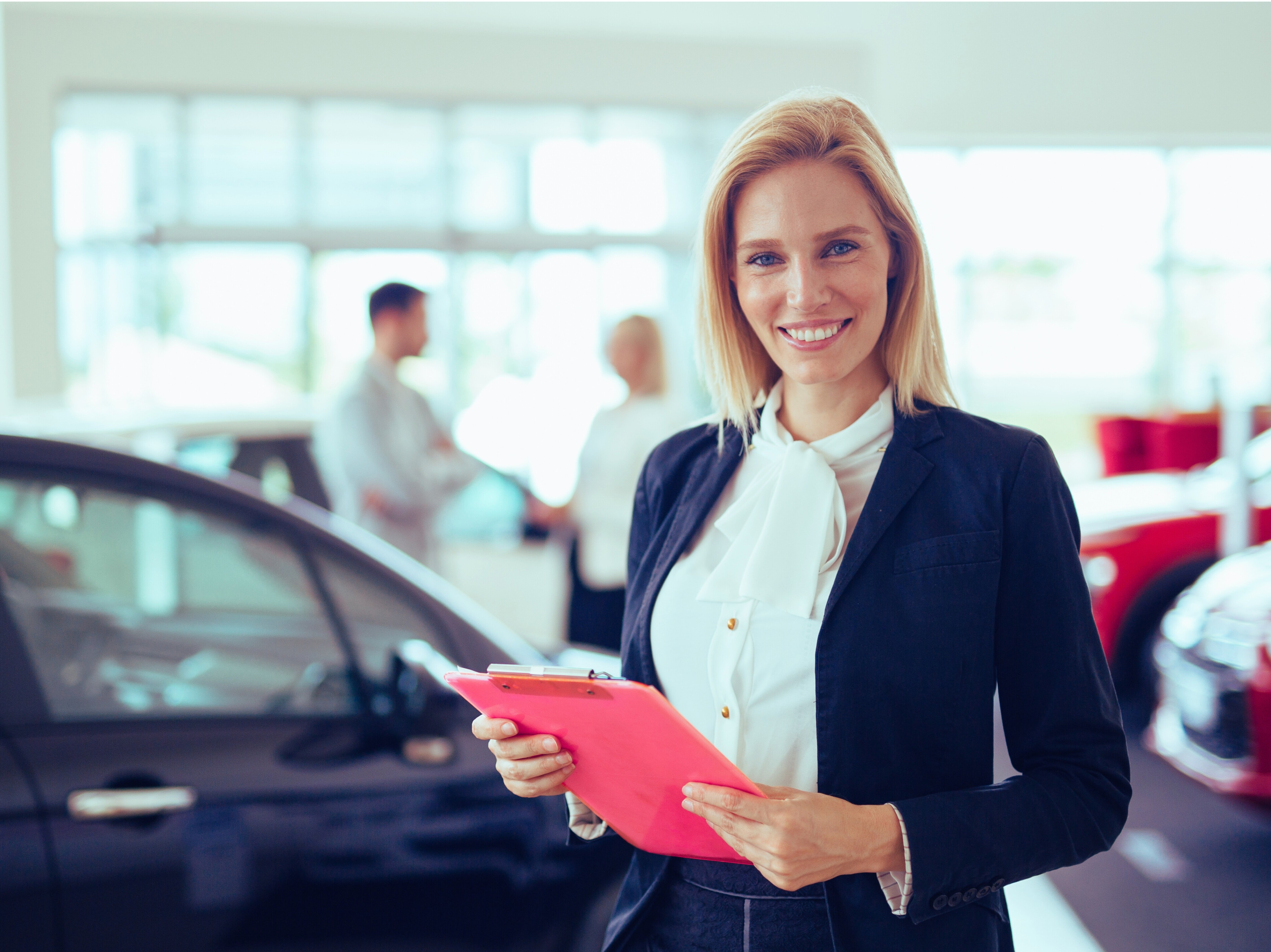 Blonde Verkäuferin steht lächeln vor einem Fahrzeug im Autohaus